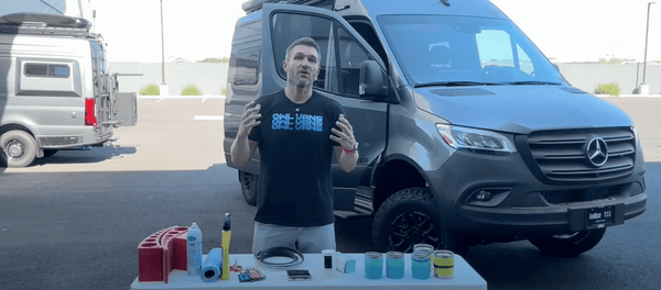 Man demonstrating Sprinter tips and tricks with a black Mercedes Sprinter van and various van accessories on a table outdoors.