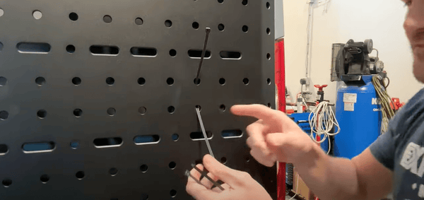 Man demonstrating zip tie trick for threading zip tie through holes on black metal van panel in workshop