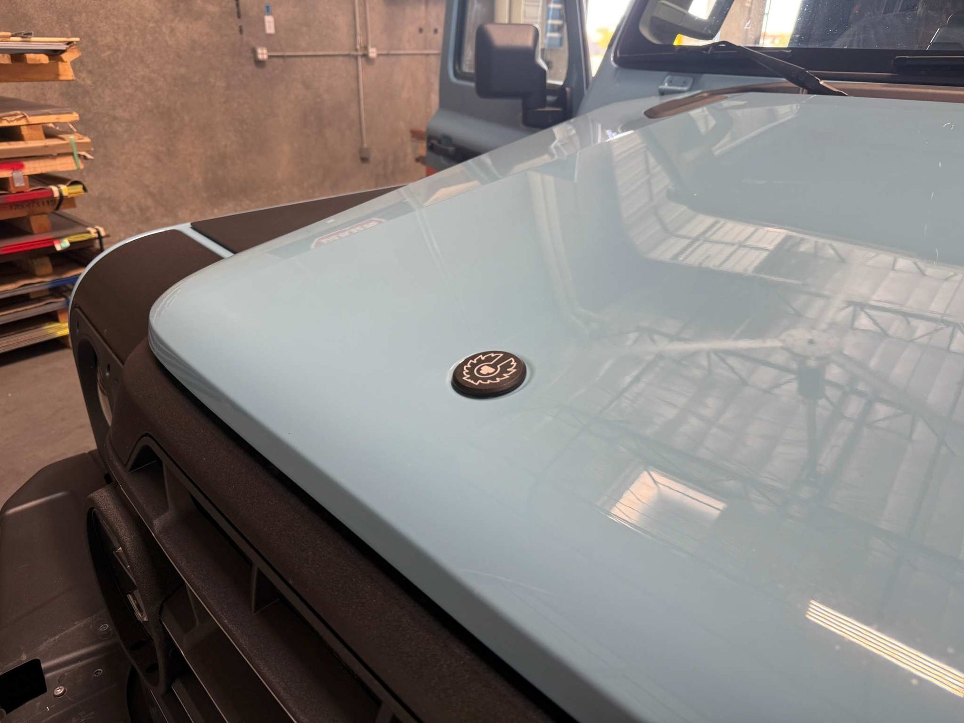 Black billet aluminum owl hood badge installed on light blue INEOS Grenadier vehicle hood in a garage setting