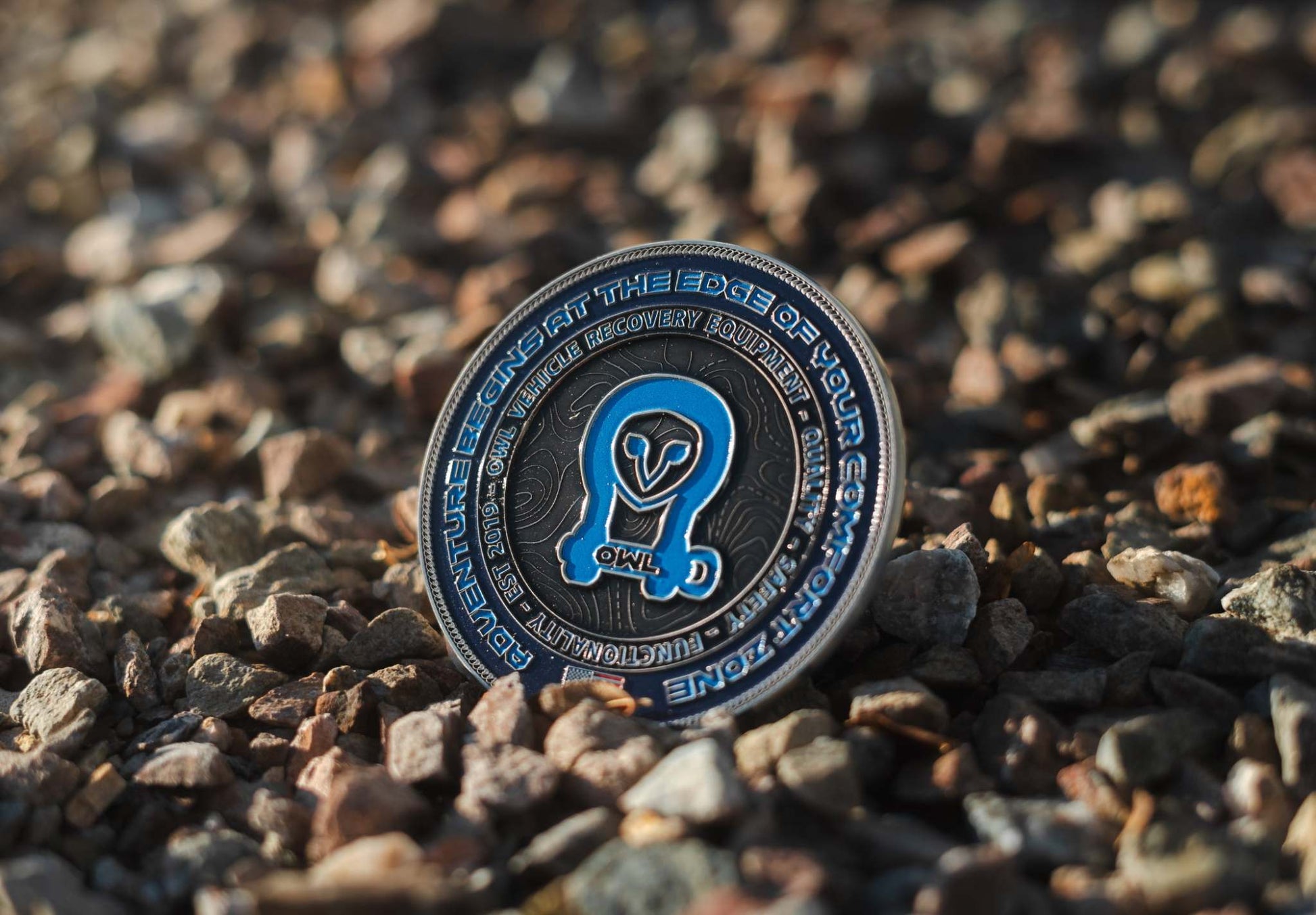 Round Owl Offroad branded challenge coin on rocky ground with blue and black design representing vehicle recovery equipment