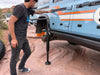 Man using Mojab hydraulic jack with mounts to lift blue INEOS Grenadier on rocky terrain