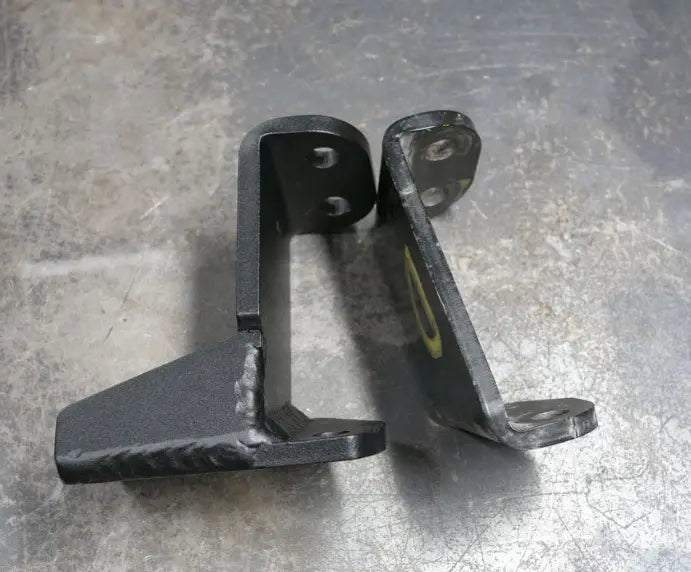 Pair of black metal Rally Strut Bump Stop support brackets for AWD vans placed on concrete floor.