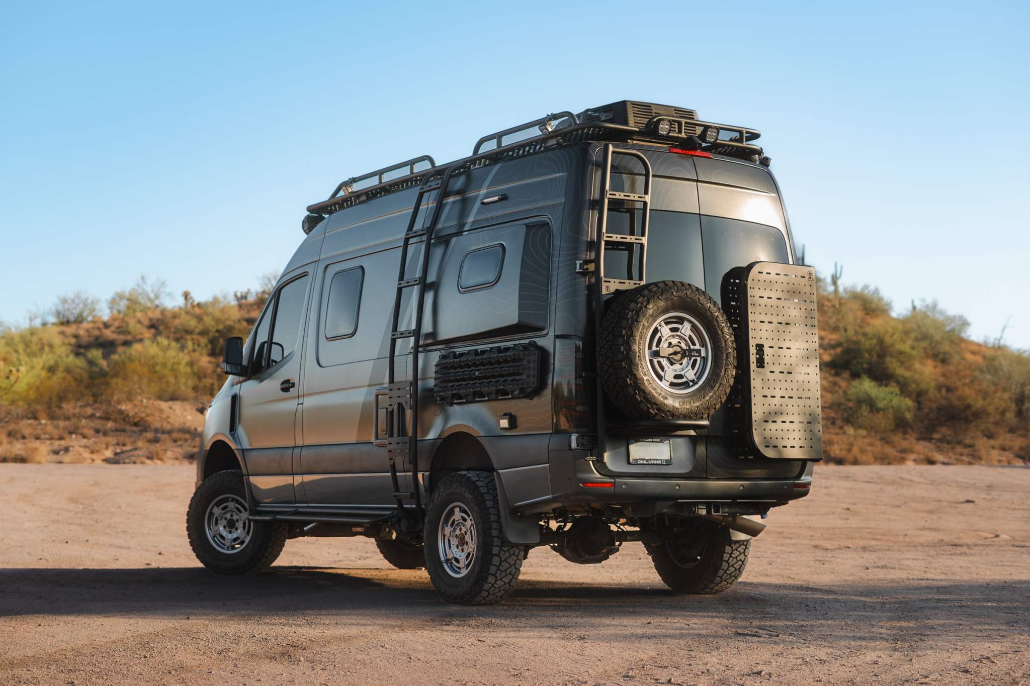 Rear view of a black Sprinter van equipped with a Rear Door B2 Classic Sprinter Bike Rack and Box Carrier mounted on the rear door, showcasing a spare tire and cargo box in a desert landscape.