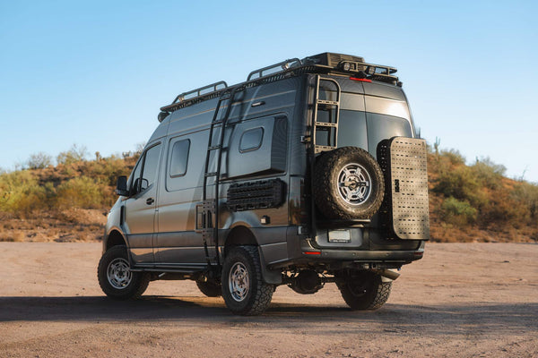 Rear view of a black Sprinter van equipped with a Rear Door B2 Classic Sprinter Bike Rack and Box Carrier mounted on the rear door, showcasing a spare tire and cargo box in a desert landscape.