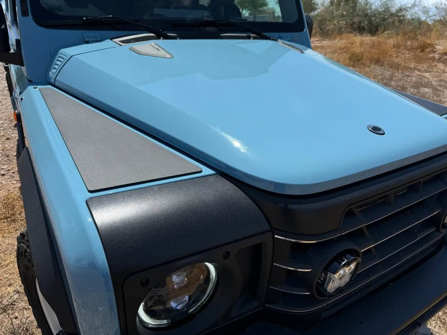 Close-up of INEOS Grenadier front with installed Grenadier Fender Armor Plates in matte black finish on a light blue vehicle.