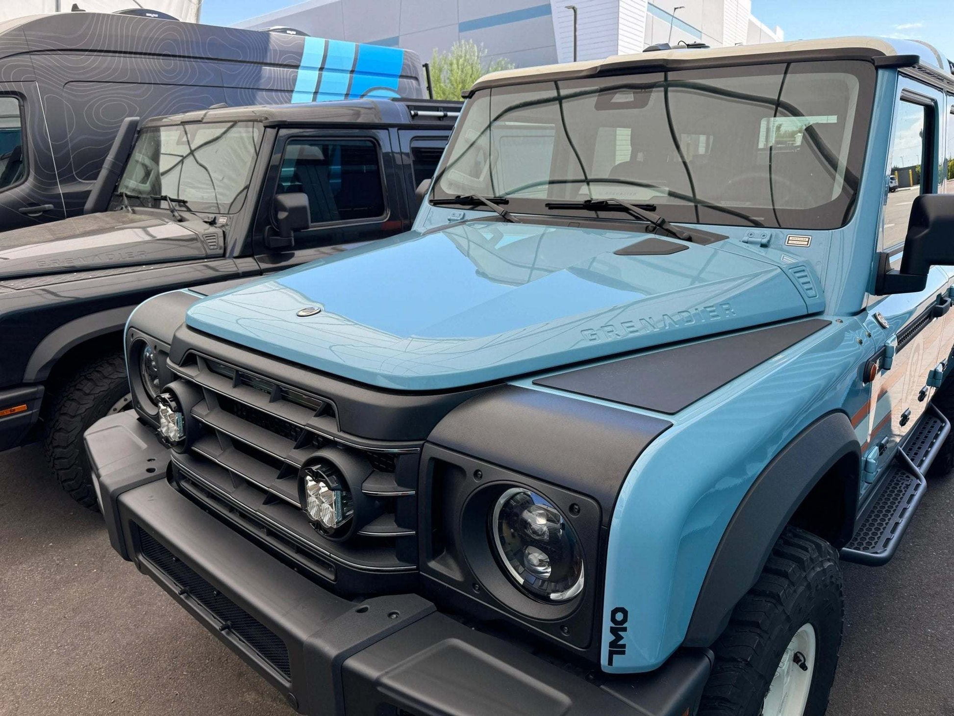Blue INEOS Grenadier with black Grenadier Fender Armor Plates on fenders and hood edges parked outdoors next to another vehicle