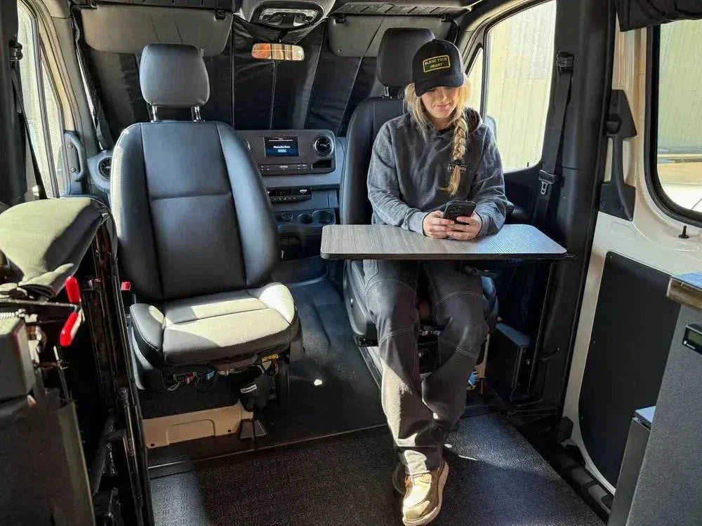 Interior view of a Sprinter van featuring a Vannon B-Pillar Mount with a fold-out table and a person seated using a phone, showcasing durable and functional overland setup equipment.
