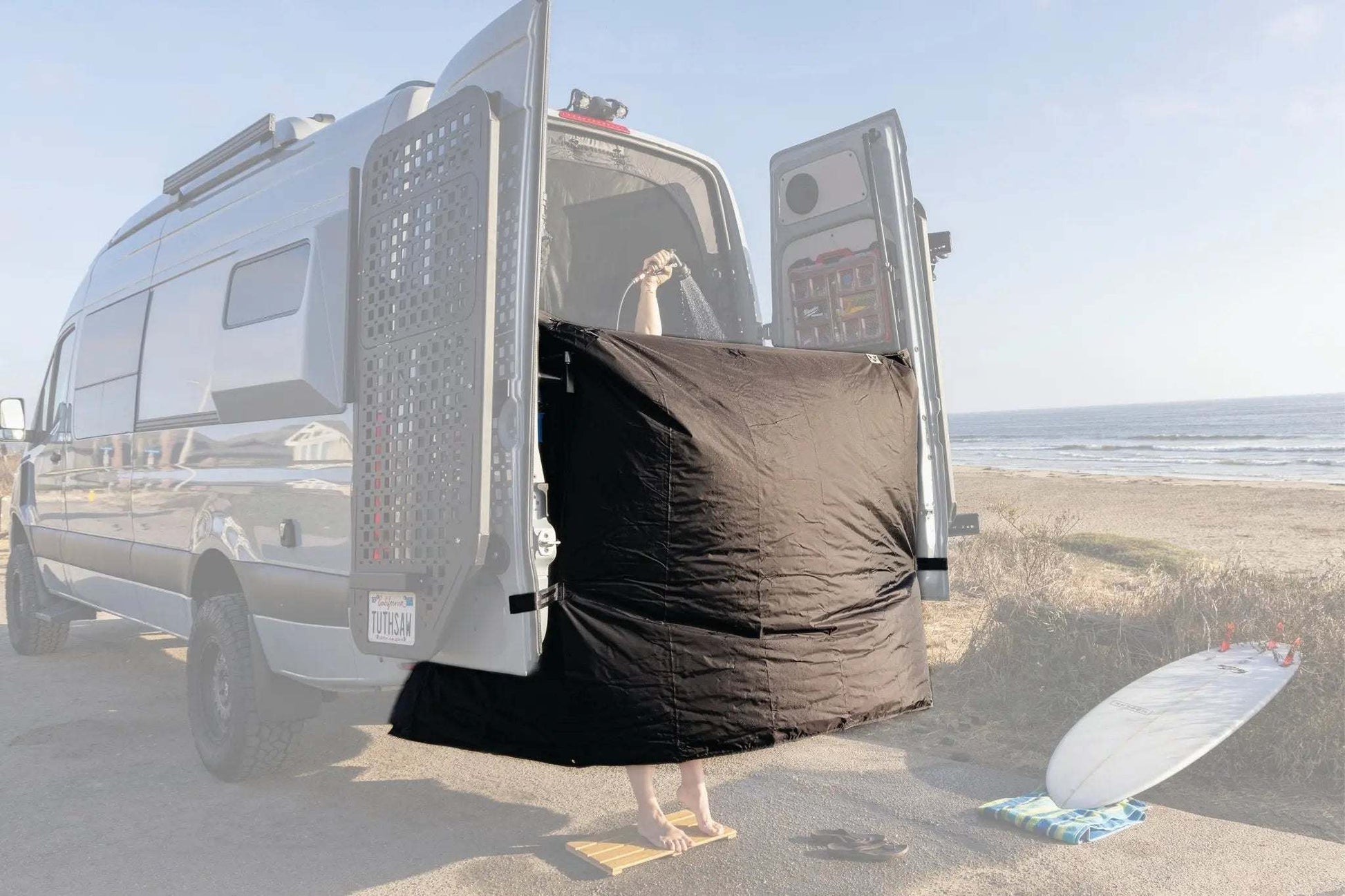 Vanlife shower curtain providing privacy and splash protection, hanging on van rear doors at a beach.