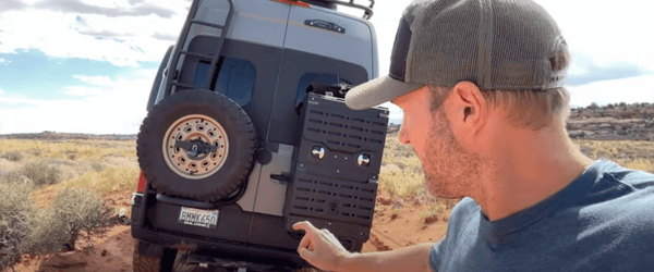 Man pointing to a tire carrier mounted on the back door of a Sprinter adventure van in a desert trail setting.