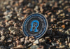 Round Owl Offroad branded challenge coin on rocky ground with blue and black design representing vehicle recovery equipment