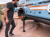 Man using Mojab hydraulic jack with mounts to lift blue INEOS Grenadier on rocky terrain
