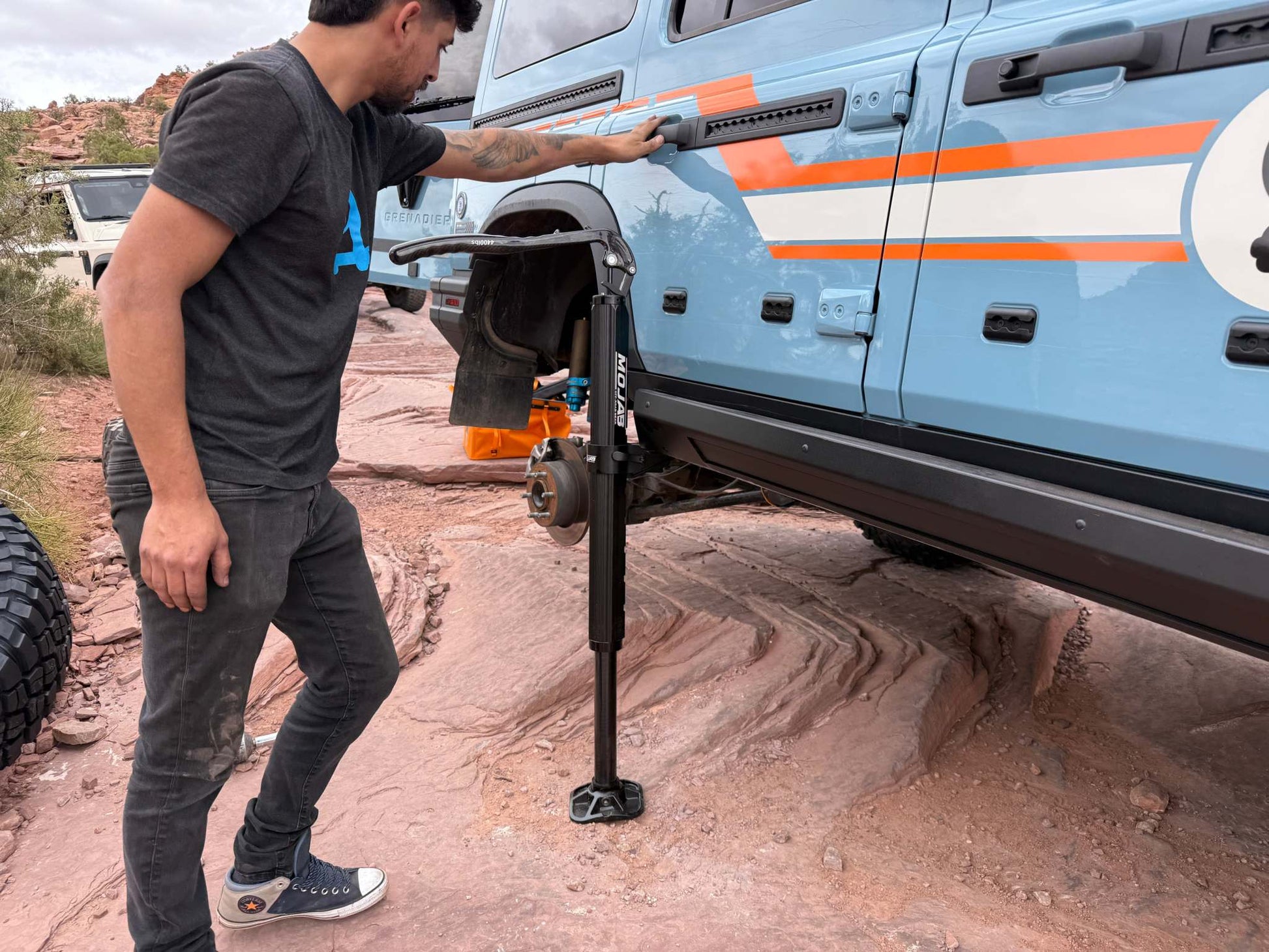 Man using Mojab hydraulic jack with mounts to lift blue INEOS Grenadier on rocky terrain