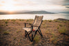 Tan ergonomic camp chair with cup holder set on rocky terrain by lake at sunset