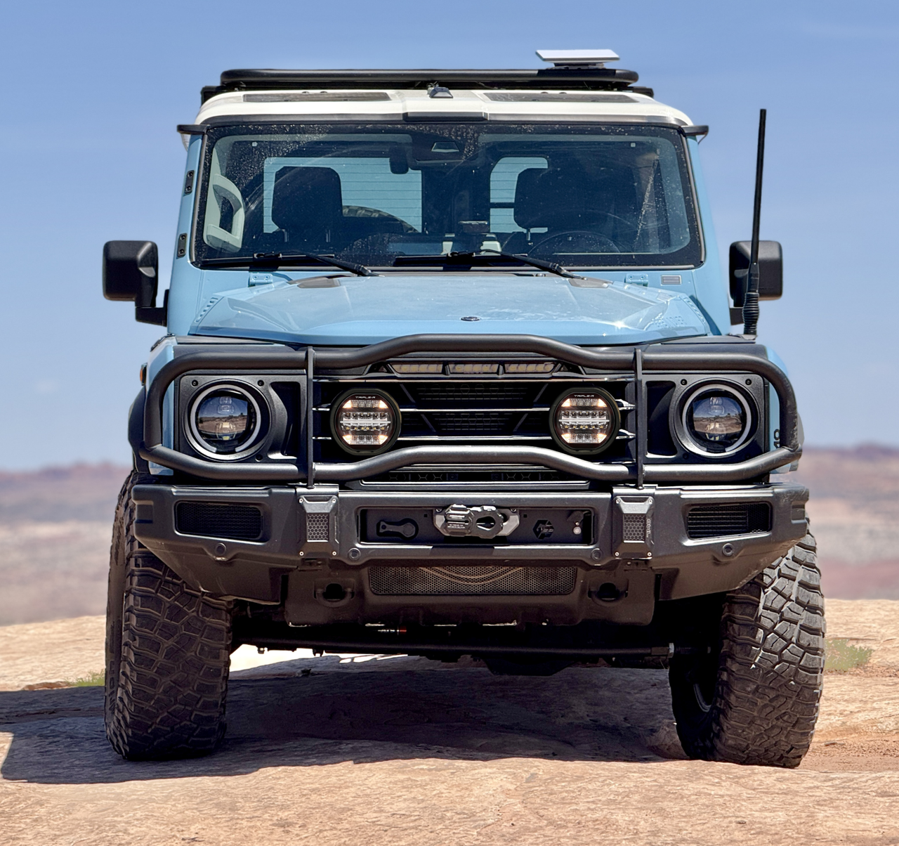 Front view of blue INEOS Grenadier equipped with black heavy-duty Grenadier Bullbar and roof rack on rocky terrain