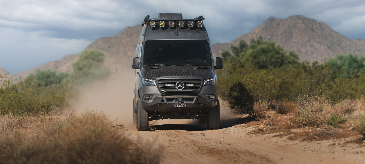 Front view of a Mercedes Sprinter van equipped with an Adax Pro Sprinter bumper driving on a dirt road in a mountainous