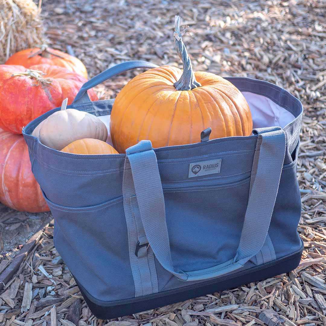 Large gear tote with pumpkins, featuring reinforced walls and multiple handles, outdoors on a wood chip surface.
