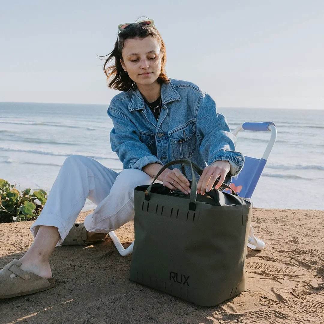 25L Soft Cooler Bag by RUX on a beach with a person sitting beside it.