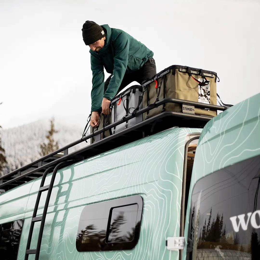 Man using Tie-Down Straps to secure gear on a rugged outdoor vehicle roof rack in a mountainous setting.