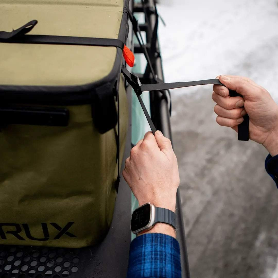 Person securing a green RUX bag with black durable tie-down straps on a vehicle rack